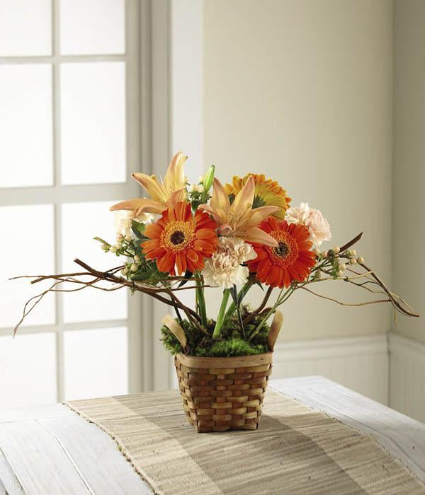  Orange gerbera daisies, orange Asiatic Lilies, and peach carnations are highlighted by peach hypericum berries, and curly willow tips, arranged to perfection in a dark stained woodchip basket with the look that they are sprouting from green moss found at the base of the display.