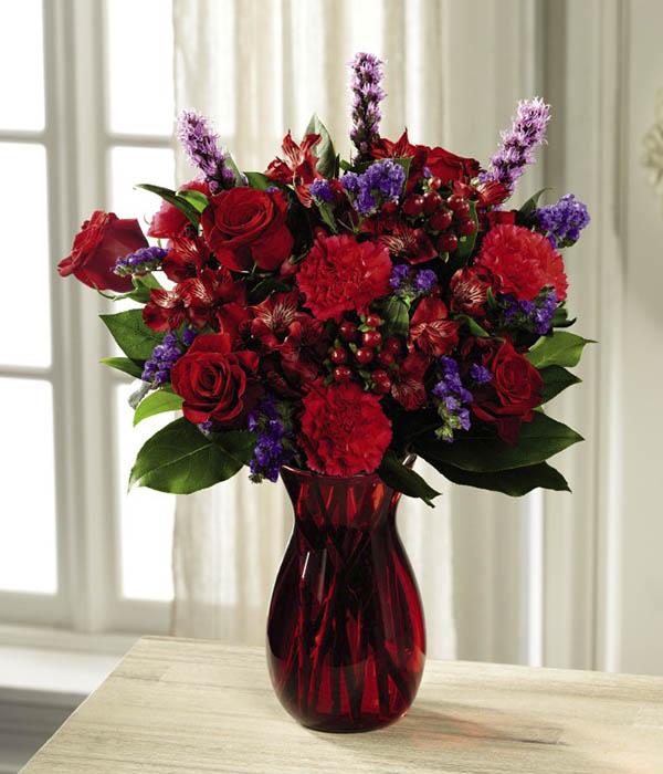 Rich red roses mingle with red Peruvian Lilies, hot pink carnations, purple statice, red hypericum berries, liatris, and lush greens, elegantly arranged in ruby red glass vase