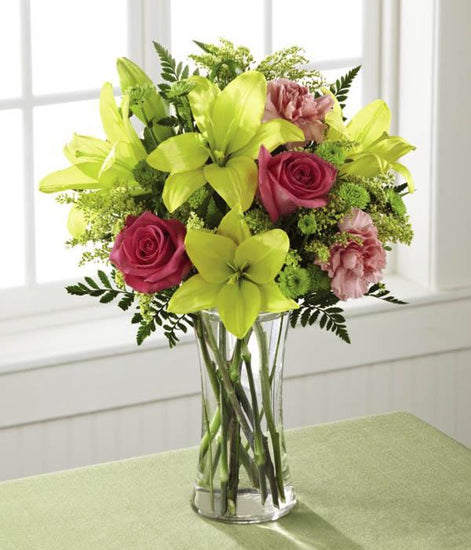  Brilliant yellow Asiatic Lilies are surrounded by hot pink roses, pink carnations, yellow solidago, and lush greens, beautifully arranged in a classic clear glass vase