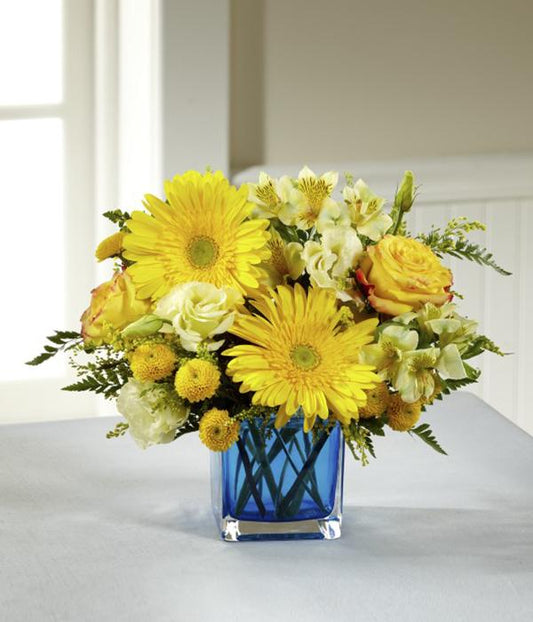 a collection of yellow blooms, including roses, gerbera daisies, Peruvian Lilies, double lisianthus, button poms, and solidago. Accented with lush greens and arranged perfectly in a modern blue glass cubed vase, standard 