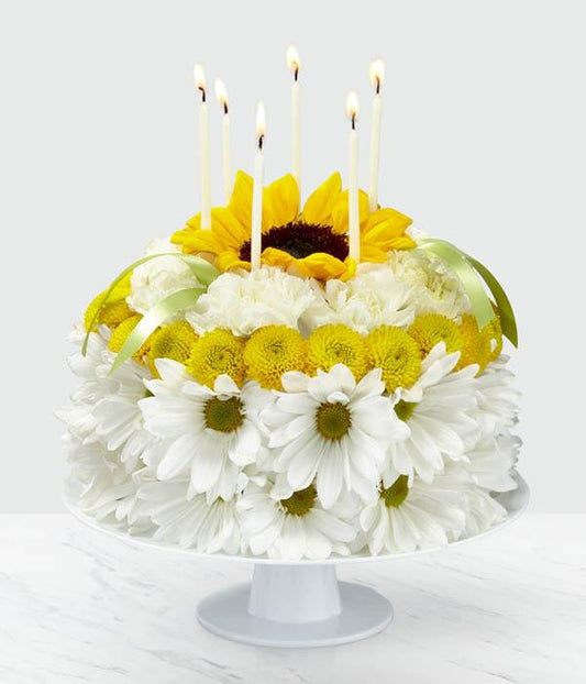 our Birthday Smiles Floral Cake IS A one-of-a-kind arrangement is frosted with a collection of bright sunflowers, yellow button pompons, white daisy pompons and white carnations. Topped with birthday candles