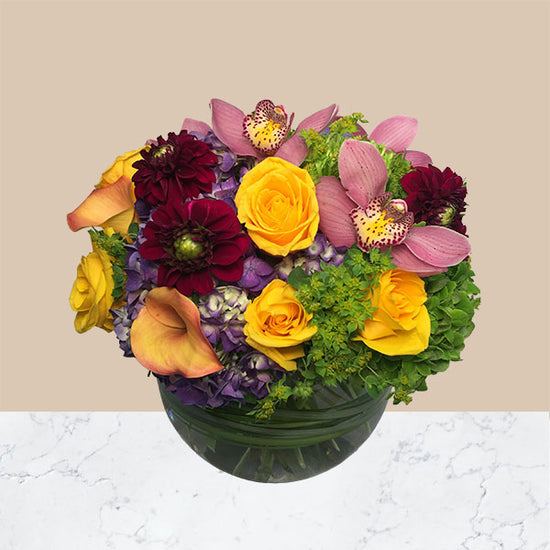 A beautiful mix of hydrangeas, roses, dahlias, cymbidium blooms and other spring flowers & lush greens presented in a clear glass bubble bowl