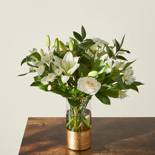 This all white arrangement features lillies,  button poms,  roses, and alstromerias in a clear vase with a gold bottom.