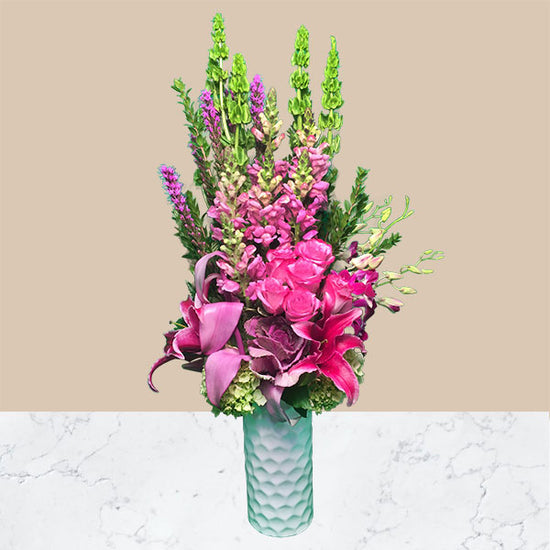 PINK AND PURPLE BLOOMS OF ROSES, STARGAZER LILIES, ANTHERIUMS, SNAPDRAGONS, DENDRO ORCHIDS, AND GREEN BELLS OF IRELAND IN A TALL WHITE CYCLINDER CERAMIC VASE