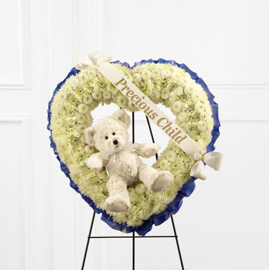 White chrysanthemums and white button poms form a gorgeous white heart-shaped wreath lined around the outside with a bright blue ribbon. Accented with an ivory plush bear at the center and a white banner that reads, "Precious Child," in gold metallic lettering