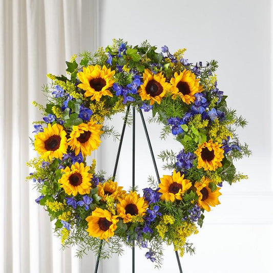 blue delpihinium, sunflowers, solidago, and lush greens arranged in a funeral wreath. Wreath is approximately 26" in diameter