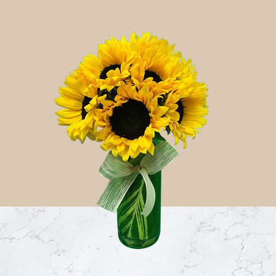 This bouquet includes assorted sunflowers arranged pave style with a ti leaf wrap in a glass cylinder vase. 